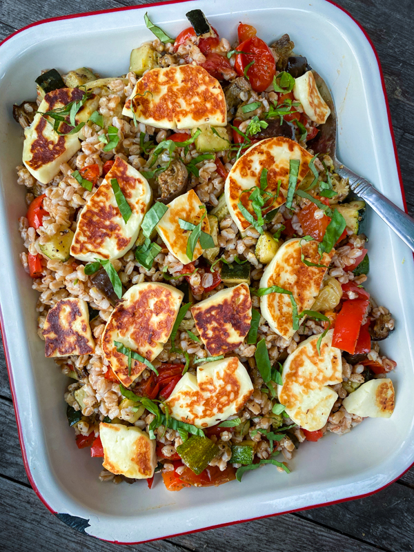 Farro with roasted vegetables and halloumi in a white and red enamel serving dish with a silver spoon.