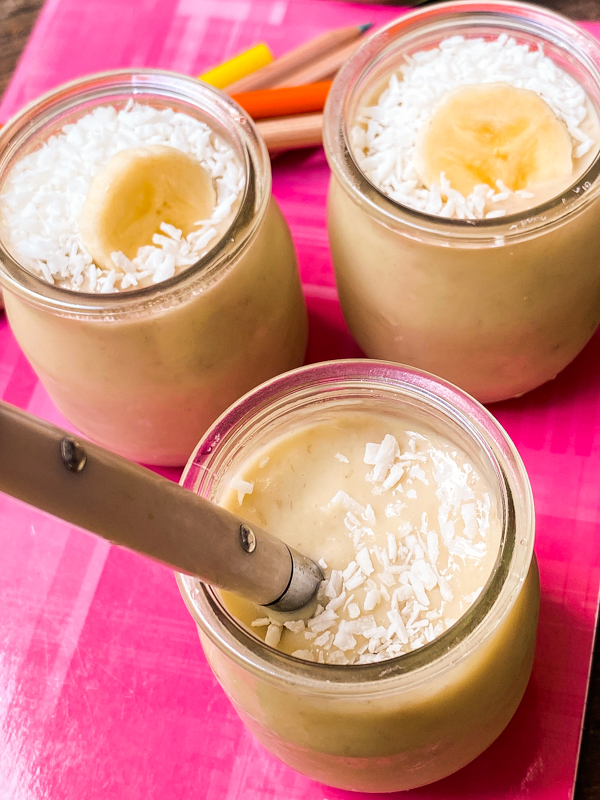 Three banana and coconut puddings in small glass jars on a notebook with coloured pencils.
