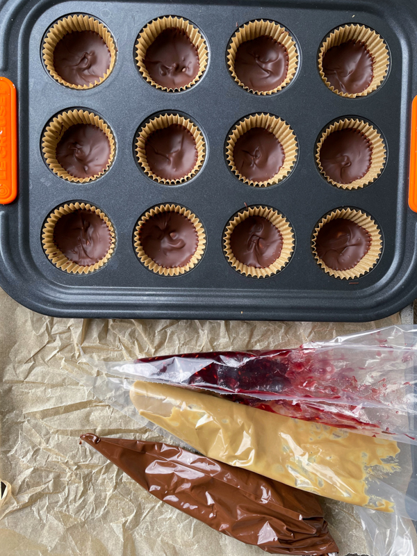 Nut free PB and J cups in progress in a mini muffin tin with piping bags of chocolate, sunbutter and jam on a baking tray.