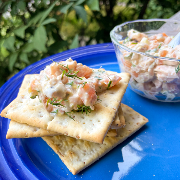 Honey-Mustard Salmon Rillettes in a small glass bowl next to a stack of crackers on a blue plate on a wooden railing.