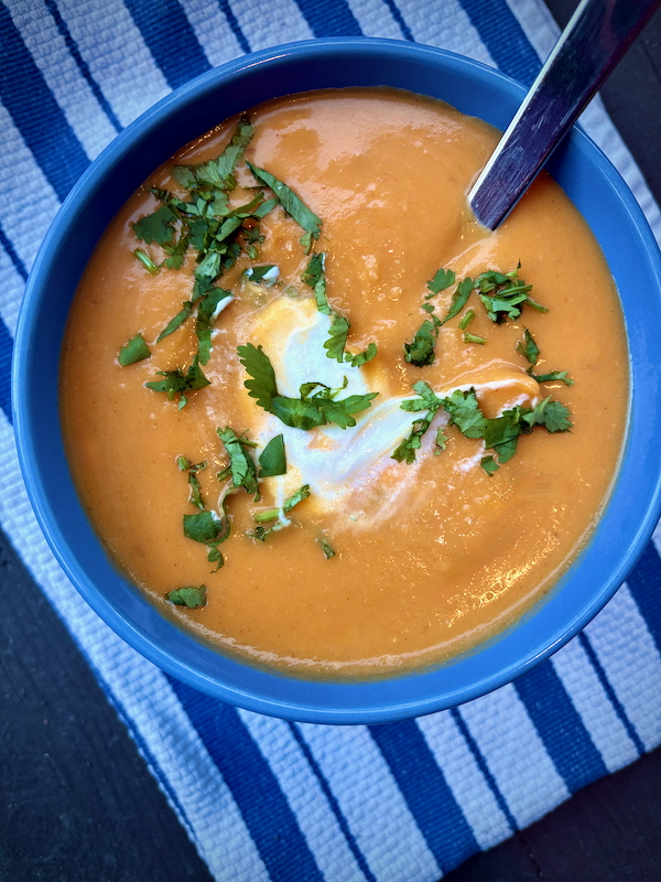A blue bowl of Coconut Curry Sweet Potato Soup with a swirl of sour cream and a sprinkle of cilantro on a blue and white napkin.