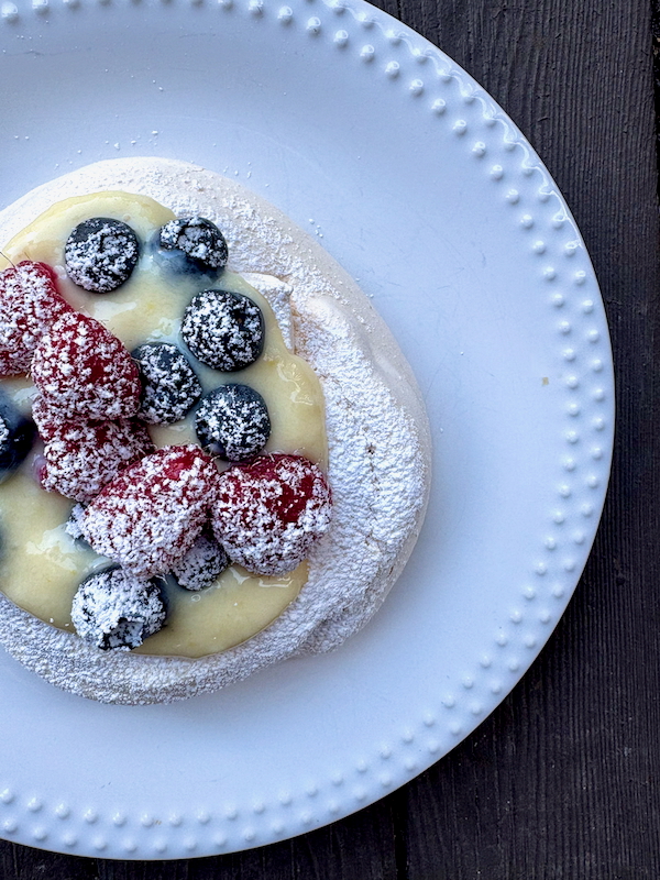 Pancake topped with blueberries and raspberries, dusted with powdered sugar on a white plate with a dotted rim. A light custard or sauce sits beneath the berries.