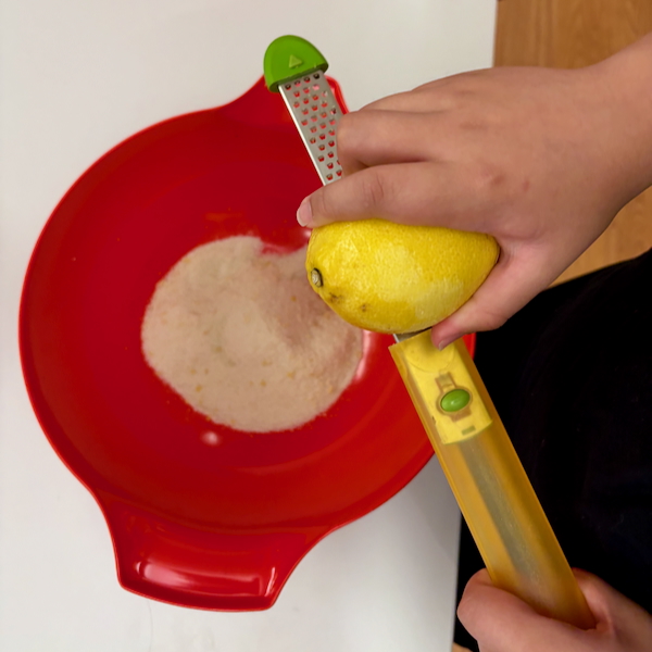 Child's hand zesting a lemon over a red mixing bowl using a small grater.