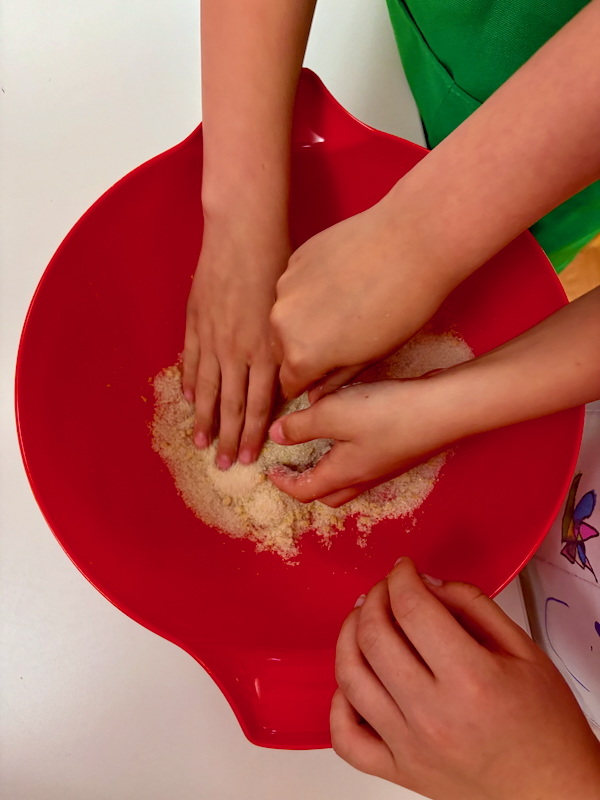 Several hands mix flour in a bright red mixing bowl on a white surface, likely a group cooking activity
