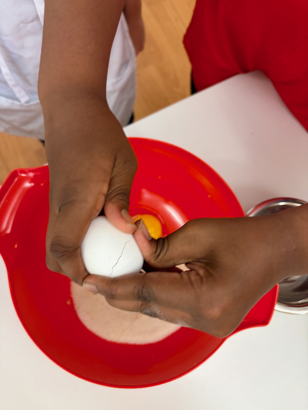 Two hands crack an egg above a red bowl, with flour visible in the bowl and a yellow yolk starting to spill in.