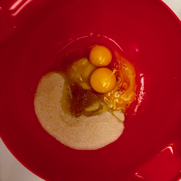 Two raw eggs with intact yolks in a red mixing bowl, surrounded by granulated sugar, ready for whisking.