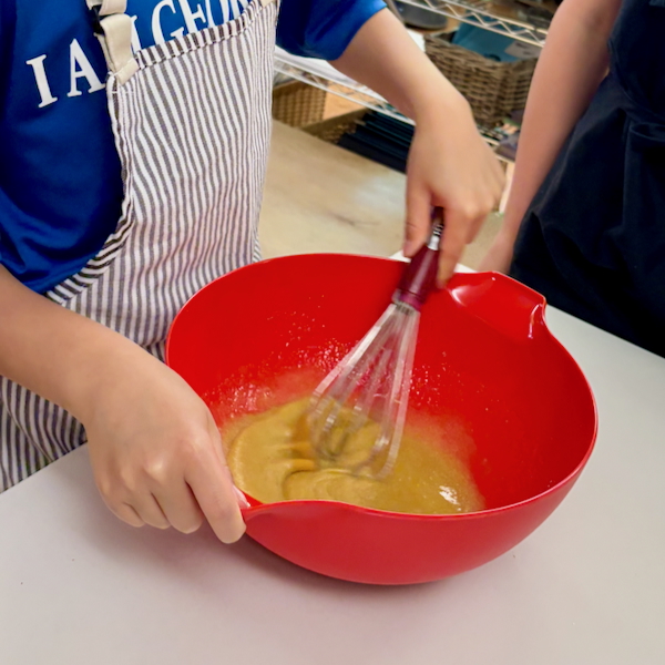 Person whisking yellow batter in a red mixing bowl on a white countertop, wearing a striped apron and blue shirt