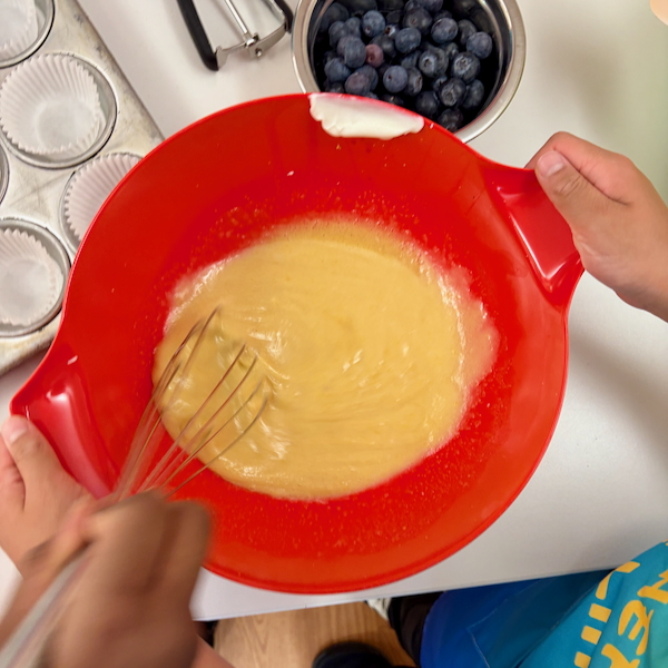 Red mixing bowl with pale batter being whisked; muffin liners and blueberries nearby.
