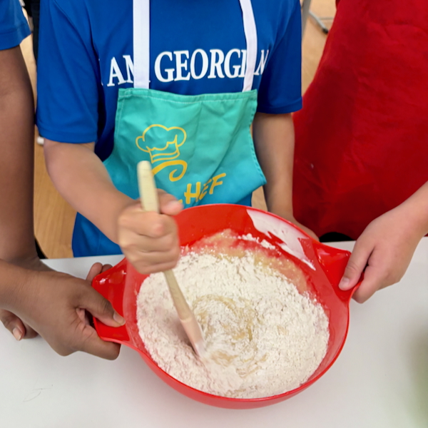 Children mixing flour in a red bowl during a baking activity, using a wooden spatula; blue apron with a chef logo visible in the scene.