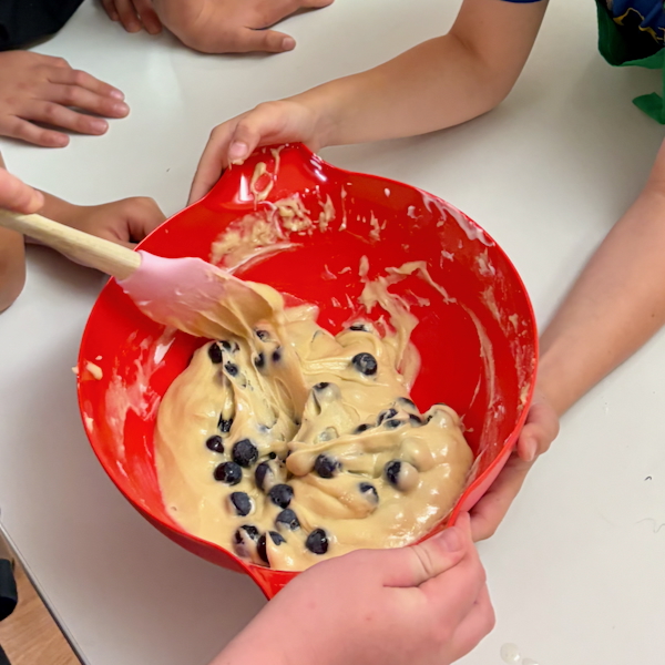 Several children’s hands hold a red mixing bowl as batter with blueberries is stirred with a pink spatula.