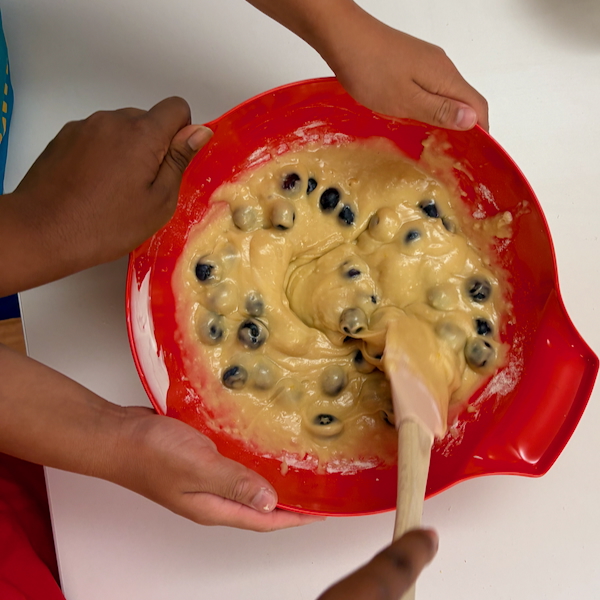 Kids' hands mix blueberry batter in a red mixing bowl using a wooden spoon, preparing dough.