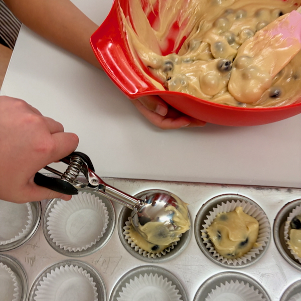Person scooping blueberry muffin batter into lined muffin cups from a red mixing bowl.