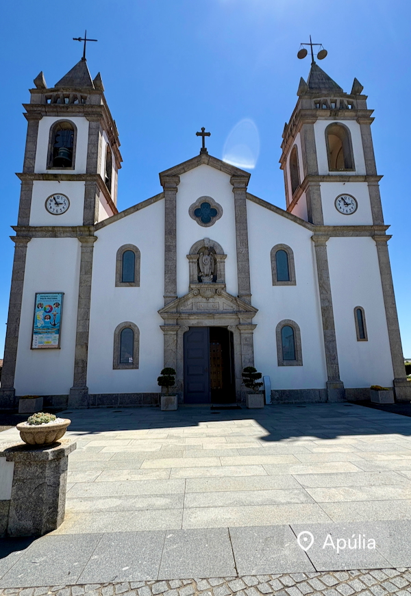 Front view of a white church with two tall bell towers, clocks, a central niche statue, and a blue door on a sunny day.