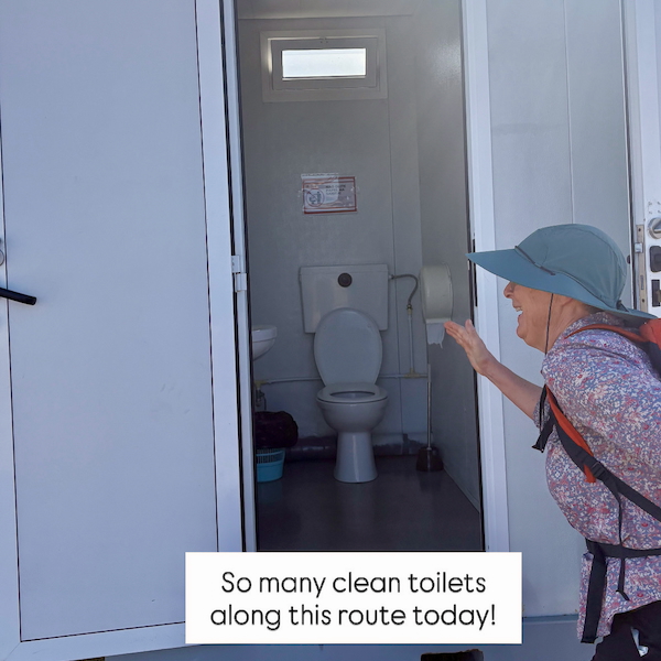 Older woman wearing a wide hat and backpack points toward an open portable toilet trailer with a white toilet inside, camera angle from outside door.