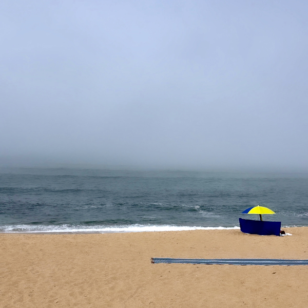 Sandy beach with a blue windbreak and a yellow umbrella near the water, a calm, foggy horizon ahead.