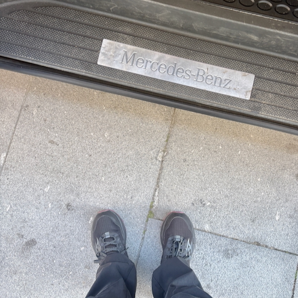 Top-down view of a person wearing black sneakers standing on gray pavement next to a Mercedes-Benz door sill plate labeled 'Mercedes-Benz'.
