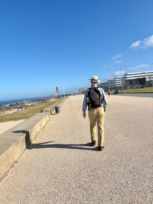 Man wearing a hat and backpack walking along a sunny coastal promenade by the sea, with modern buildings in the distance.