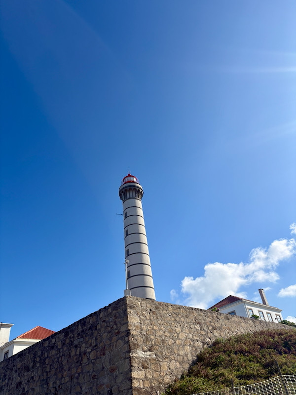 Tall white lighthouse with a red top rises above a rugged stone seawall under a clear blue sky.