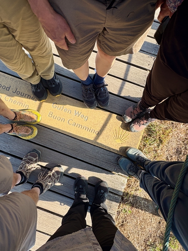 Group of hikers standing in a circle on a wooden boardwalk, feet around a tile engraved with 'Buen Camino' in several languages.