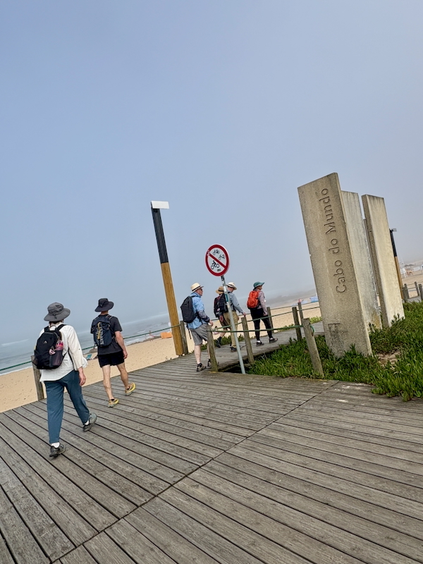 People walk along a wooden boardwalk toward a sandy beach with the ocean on the left; a red no-bicycles sign stands near angled concrete posts reading Cabo do Mundo