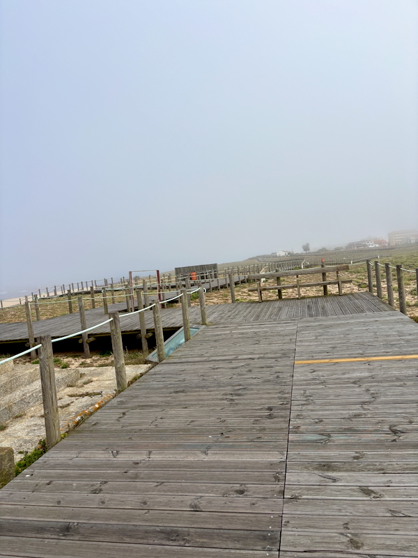 Wooden boardwalk along a fenced coastal path fading into thick fog.