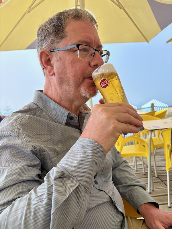 Man with gray hair and glasses sipping a pale beer from a tall glass labeled Super Bock at an outdoor patio with yellow chairs and umbrellas.