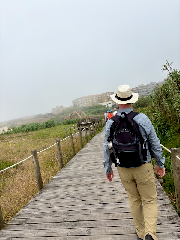 Person with backpack and straw hat walking along a wooden boardwalk beside grassy dunes toward distant buildings by the coast.