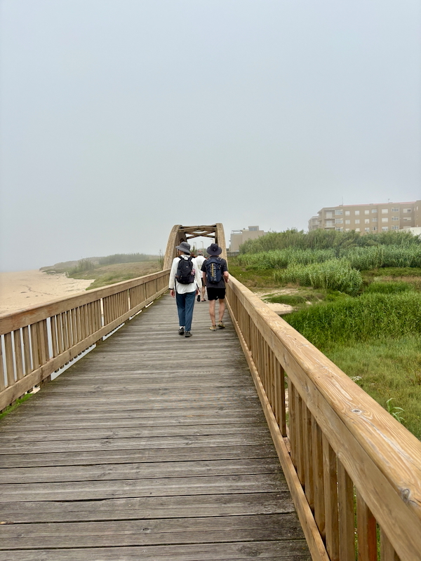 Two hikers with backpacks walk away on a wooden boardwalk toward a foggy beach.