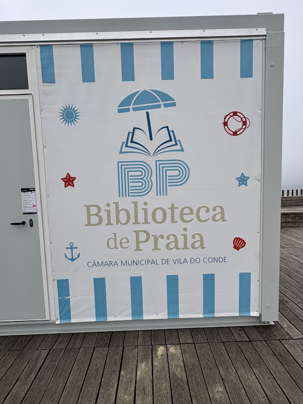 Sign for Biblioteca de Praia (Beach Library) with BP logo and sea-themed icons on a portable panel on a wooden boardwalk, Vila do Conde.