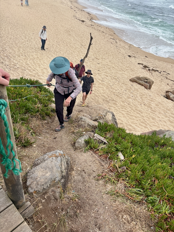 Hikers ascend a rocky coastal path from the sandy beach toward the water, using a rope handrail on the left.