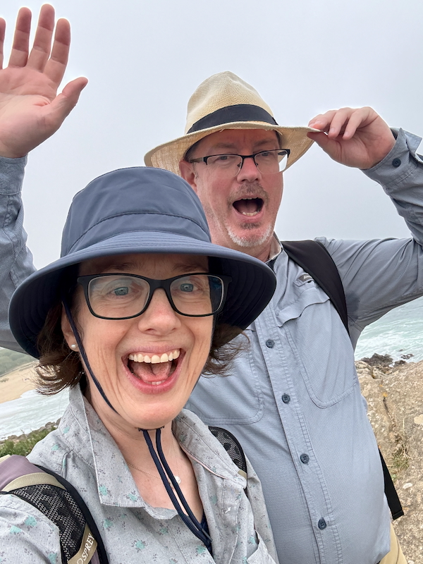 Two friends take a cheerful selfie on a rocky coastal overlook, wearing hats and sunglasses with the ocean in the background.