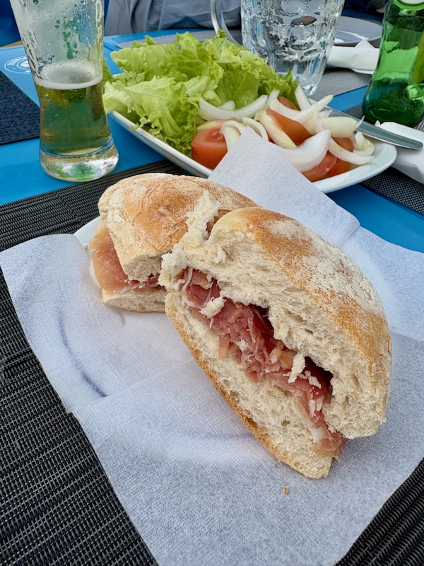 Crusty ham sandwich cut in half on a white napkin, with a bite taken from one half on a blue table.