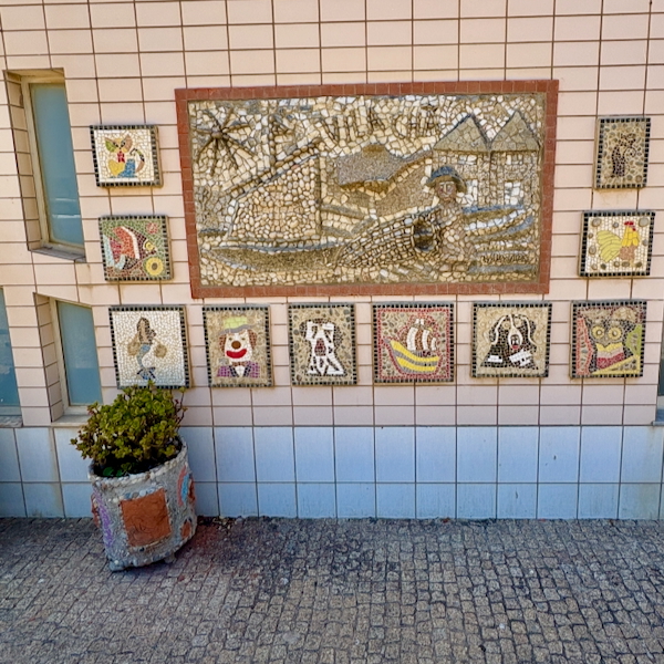 Exterior wall mosaic: large central tile mural with smaller surrounding mosaic pictures and a potted plant on the left, on tiled masonry.