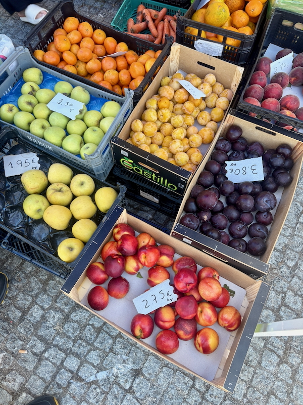 Crates of fresh fruit at a market: green and yellow apples, peaches, plums, and citrus with handwritten price tags.