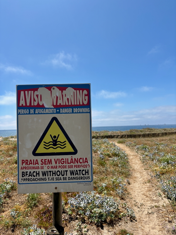 Warning sign at a coastal beach warning about drowning danger with a person in water icon and 'Beach Without Watch' text in English and Portuguese.