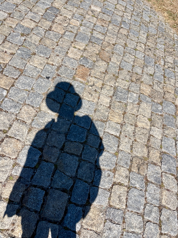 Shadow of a person wearing a hat cast on a cobblestone pavement.