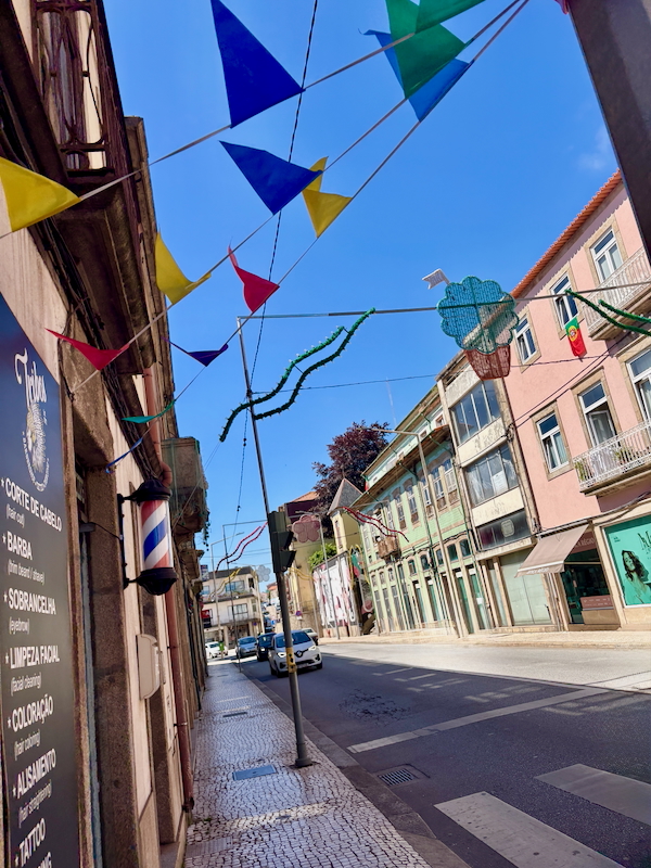 Colorful triangular bunting hangs over a sunlit street lined with pastel storefronts and a barber pole on the left, under a blue sky.