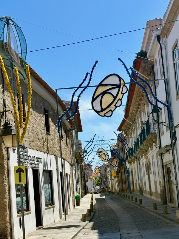 Narrow cobblestone street lined with historic buildings and decorative festival lights arching overhead, under a blue sky.