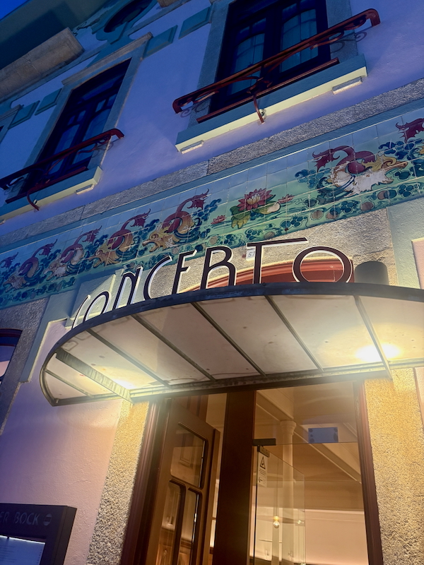 Exterior entrance with curved glass canopy and glass doors; blue building with a colorful floral tile frieze above.