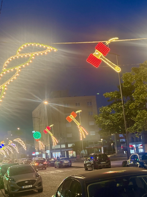 Night street scene with glowing holiday lights shaped like gift boxes and loops above a cobblestone road at dusk/dawn-tinted sky.