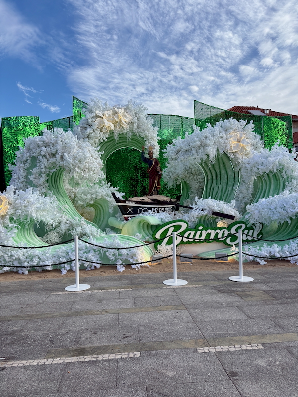 Carnival float with green wave shapes, white foam, and a statue in a boat at a public square.