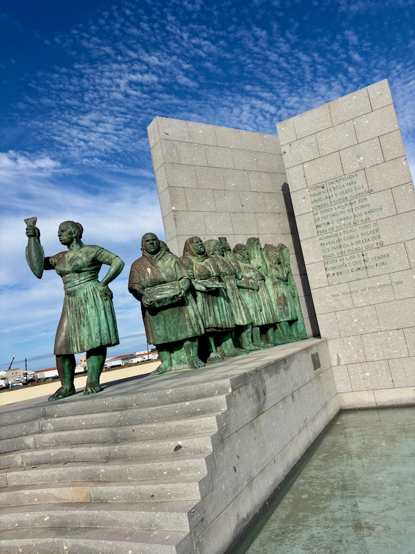 Bronze statue group of six people on a stepped stone pedestal beside a granite memorial wall under a blue sky; outdoor memorial sculpture.