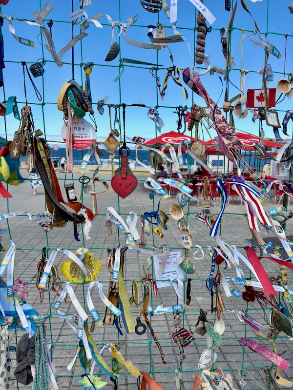 Colorful ribbons and trinkets tied to a wire fence along a beachfront boardwalk.