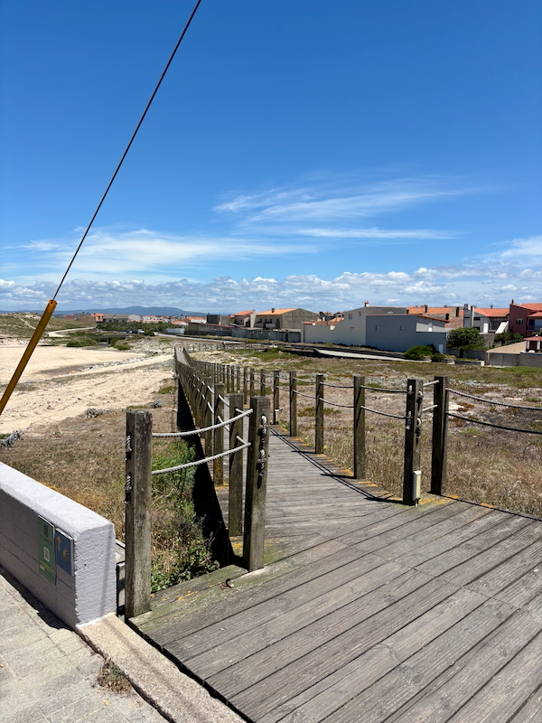 Wooden boardwalk with railings extending toward a row of houses in a coastal neighborhood under a blue sky.