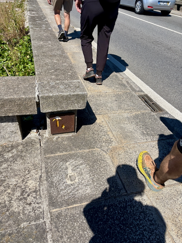 Three pedestrians walk along a sunlit stone sidewalk beside a road; a small brown metal utility box with a white hammer symbol is set into the curb near the edge of the path.
