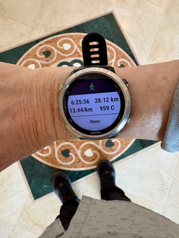 Garmin smartwatch on a wrist showing running stats: 28.12 km, pace 13:44/km, 959 kcal, with Done at bottom. Indoor tiled floor visible.