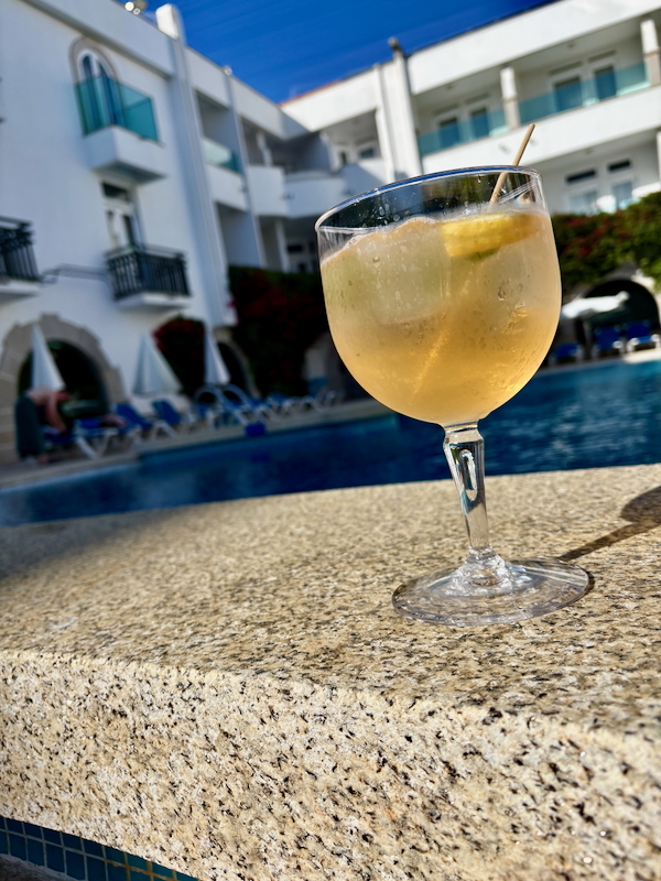 Cocktail glass with ice and a lemon wedge sitting on a poolside stone ledge at a sunny resort pool.