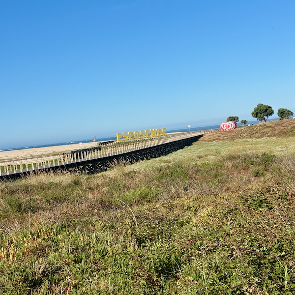 Wooden boardwalk extending to a sandy beach under a clear blue sky, with a yellow Esposende sign and a heart logo in the distance.