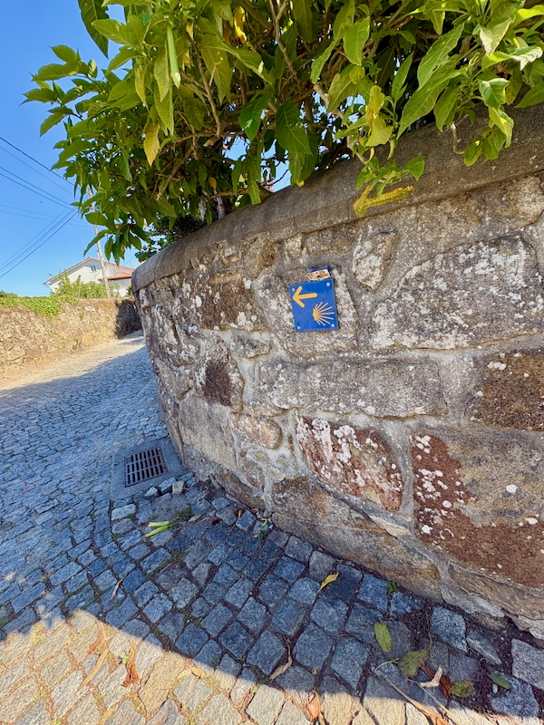 Blue square waymark with a left-pointing arrow and a shell on a curved stone wall beside a cobblestone street.
