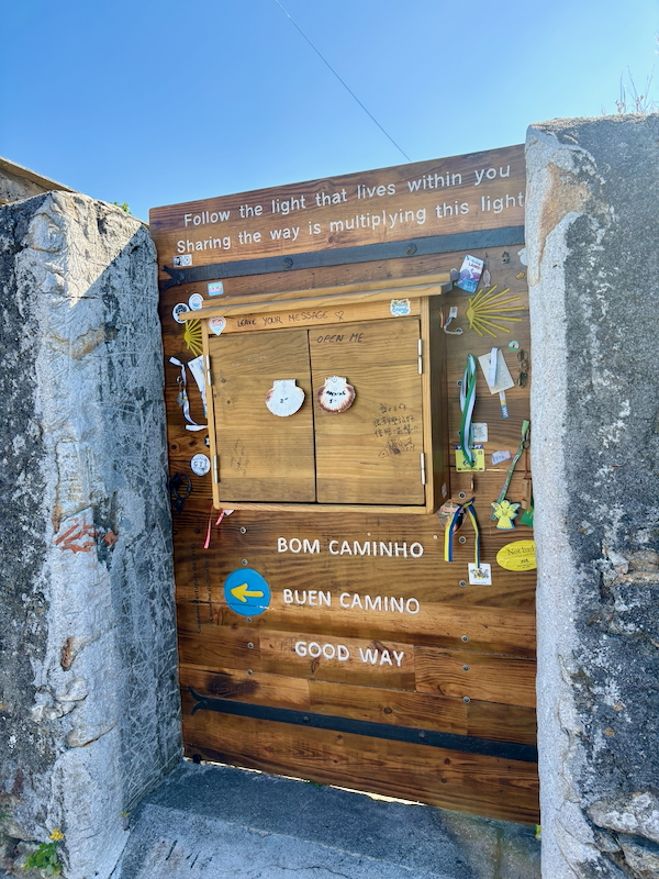 Wooden cabinet in a stone doorway with signs 'Bom Caminho', 'Buen Camino', 'Good Way' and a blue arrow, surrounded by notes and trinkets.